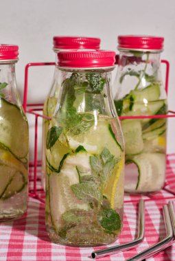 Kırmızı kapaklı cam şişeler, içinde salatalık, limon dilimleri ve nane yaprakları bulunan arınma suyu bulunan kırmızı ve beyaz kareli masa örtüsü. Metal pipetler 