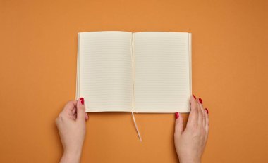 Turuncu bir arka planda, temiz ve çizgili sayfaları olan açık bir defter tutan kadın eller. Metin ya da planlama için düzen.