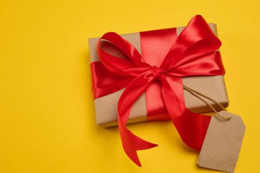 Kırmızı ipek fiyonklu bir hediye kutusu ve sarı arka planda boş bir kahverengi etiket.