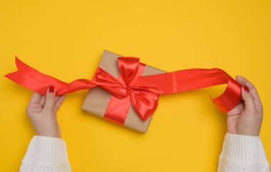 Beyaz örgü kazaklı bir kadının elleri büyük kırmızı saten bir fiyonklu hediye kutusunu tutuyor. Yukarıdan görünüm, sarı arkaplan.