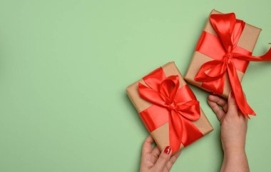 İki kadın el, yeşil arka planda kırmızı kurdeleyle bağlanmış hediye kutularını tutuyor. Boşluğu kopyala