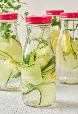Kırmızı kapaklı cam şişeler, içinde salatalık, limon dilimleri ve nane yaprakları bulunan detoks suyla dolu beyaz masa.