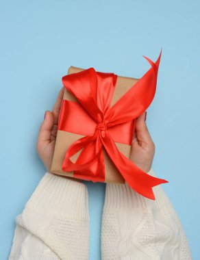 Beyaz örgü kazaklı bir kadının elleri büyük kırmızı saten bir fiyonklu kare bir hediye kutusu tutar. Yukarıdan mavi arkaplana göre görüntüle.