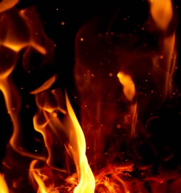 Turuncu ve sarı alevlerin yoğun dilleri, havada parlayan küçük kıvılcımlar ve kül parçacıkları, siyah arkaplan