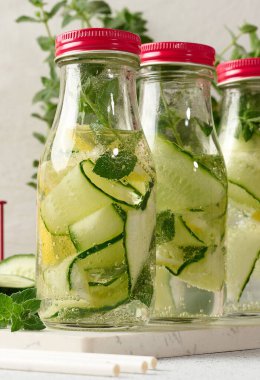 Kırmızı kapaklı cam şişeler, içinde salatalık, limon dilimleri ve nane yaprakları bulunan detoks suyla dolu beyaz masa.