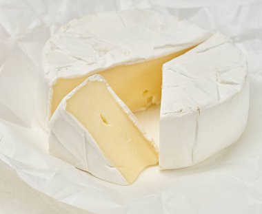 Beyaz küflü, yuvarlak bir Camembert peyniri, bir üçgen dilimlenmiş, peynir buruşmuş beyaz ambalaj kağıdının üzerinde yatıyor.