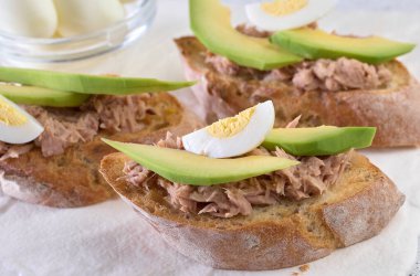 Bir dilim kızarmış ekmek, doğranmış ton balığı, olgun avokado dilimleri ve haşlanmış yumurta çeyreklikleri, beyaz masa.