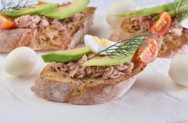 Bir dilim kızarmış ekmek, doğranmış ton balığı, olgun avokado dilimleri ve haşlanmış yumurta çeyreklikleri, beyaz masa.