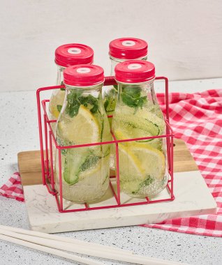 Kırmızı kapaklı cam şişeler, içinde salatalık, limon dilimleri ve nane yaprakları bulunan detoks suyla dolu beyaz masa.