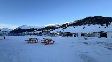 Livigno, İtalya - 8 Şubat 2026: Milano Cortina 2026 Olimpiyat sahasında kırmızı piknik masaları, yemek tezgahları ve gün batımında karlı Alp dağları ile İtalya 'nın Livigno kentinde