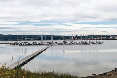 La Baie, Kanada - 19 Ağustos 2025: Arkaplanda uzak tepeler ve kıyı şeritleriyle bulutlu bir gökyüzünün altında sakin bir su üzerinde demirlemiş yelkenli tekneleri olan bir marinaya giden ahşap iskele