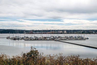 La Baie, Kanada - 19 Ağustos 2025: Arkaplanda uzak tepeler ve kıyı şeritleriyle bulutlu bir gökyüzünün altında sakin bir su üzerinde demirlemiş yelkenli tekneleri olan bir marinaya giden ahşap iskele