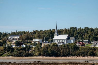 Saguenay Nehri 'nin karşısındaki panoramik manzara Baie-Sainte-Catherine köyünün kıyılarını renkli evler ve ağaçlarla gösteriyor..