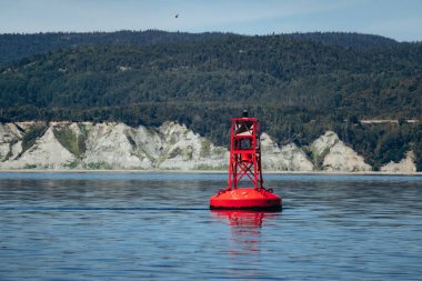 Baie Sainte Catherine yakınlarındaki St Lawrence Nehri 'nin sakin sularında yüzen kırmızı seyrüsefer şamandırası, ormanlık tepeler ve açık mavi gökyüzünün altında kıyı kayalıkları