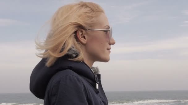 Усміхнена дівчина в сонцезахисних окулярах, що йдуть вітряним днем біля пляжу — стокове відео