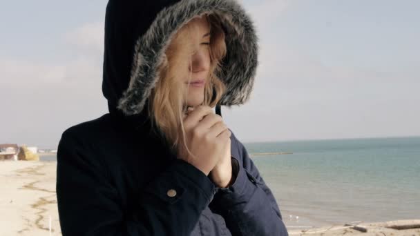 Молодая блондинка в пуховой куртке купается на солнце на пляже и улыбается — стоковое видео