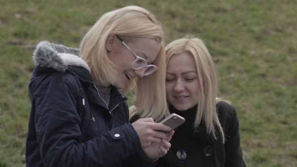 Dos chicas sentadas en el banco en el parque de la ciudad y mirando el teléfono sonriendo — Vídeos de Stock