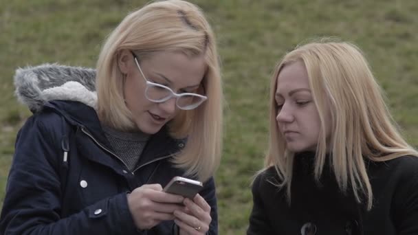 Δύο κορίτσια κάθονται στον πάγκο στο πάρκο της πόλης και να βλέπουν το τηλέφωνο χαμογελώντας — Αρχείο Βίντεο