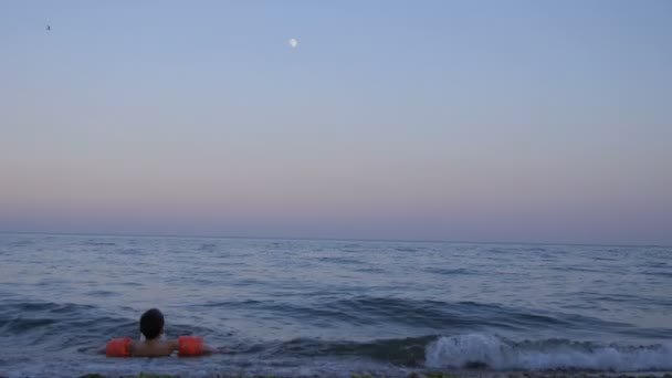 Menino brincando em ondas na praia em braçadeiras infláveis no crepúsculo — Vídeo de Stock