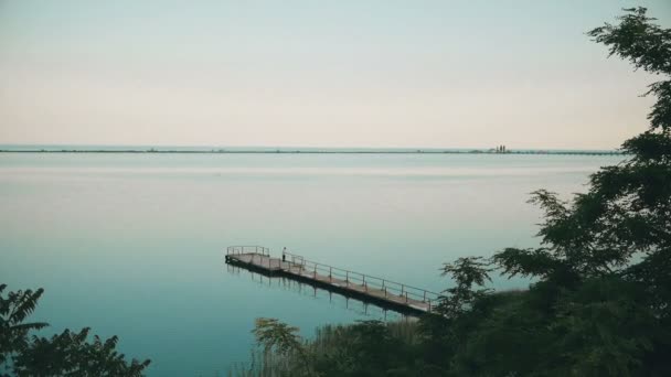 Vista del paisaje del amanecer en el lago con muelle de madera y pescador solo — Vídeos de Stock