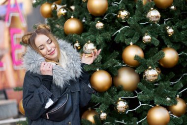 Altın topları ve ışıklarıyla Noel ağacının arka planında poz veren, gözleri kapalı, mutlu, çekici genç bir kadın..                               