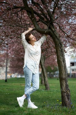 Genç, çekici, kızıl saçlı, beyaz bir bluz ve çiçek açan sakura 'nın arka planında poz veren bir kot giyen kadın..                               
