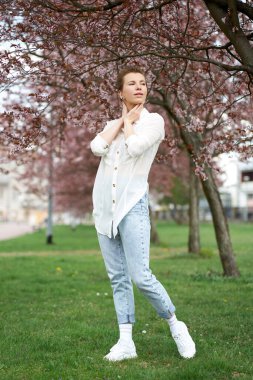 Genç, çekici, kızıl saçlı, beyaz bir bluz ve çiçek açan sakura 'nın arka planında poz veren bir kot giyen kadın..                               