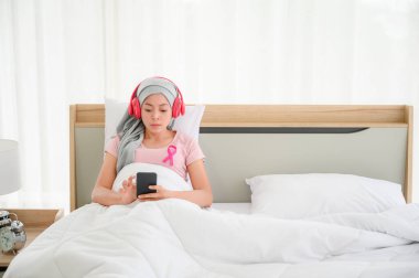 Genç Asyalı tesettürlü kadınlar yataktaki akıllı telefona oturur ve kulaklıkları takarlar. Göğüs kanseri hastalığının iyileşmesini temsil ediyor. Göğüs kanseri kavramı, kanser önleme kavramı.