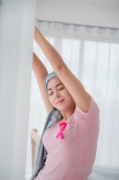 Young asian women wearing hijab Open the curtains in the bedroom to get the morning sunshine. Attaching a pink ribbon represents recovery from a breast cancer patient. Breast cancer concept, cancer pr