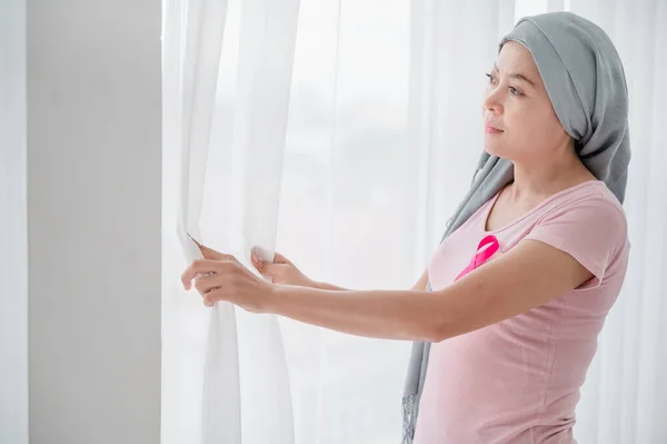 Young asian women wearing hijab Open the curtains in the bedroom to get the morning sunshine. Attaching a pink ribbon represents recovery from a breast cancer patient. Breast cancer concept