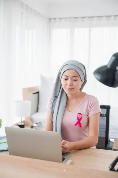 Young Asian woman wearing a hijab uses smartphone and laptop at her desk. Attaching a pink ribbon represents recovery from a breast cancer patient. Breast cancer concept, cancer prevention concept.