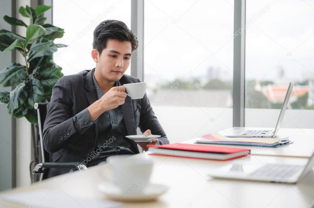 Confident and smart handsome business man in formal wear sipping coffee at his desk. With a laptop in the background. The concept of working rest in the workspace.