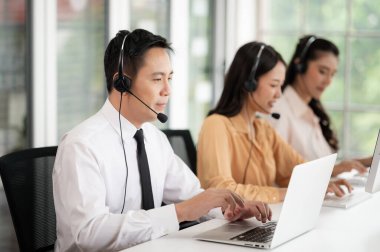 Asya 'daki çağrı merkezi personeli mikrofonlu kulaklık takıyor. Masa ve bilgisayarlarda müşterilere hizmet ederken gülümseyin. Hizmet konsepti ve danışmanlık. İletişim konsepti.