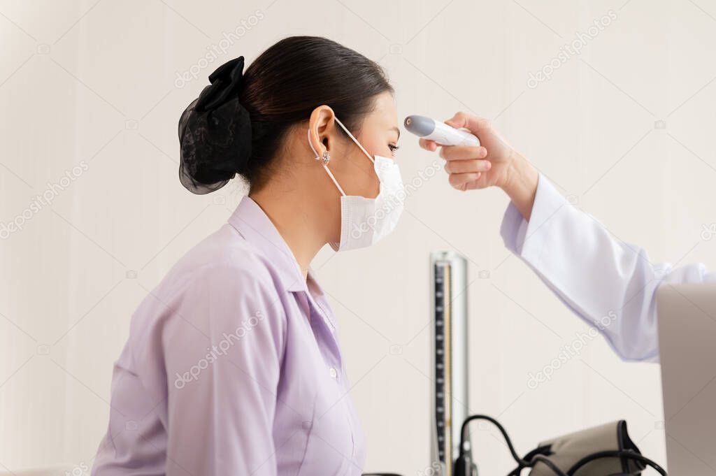 Doctor checking the temperature of a young woman patient with a digital thermometer at the auscultation room. Concept of preventing the spread of COVID-19.
