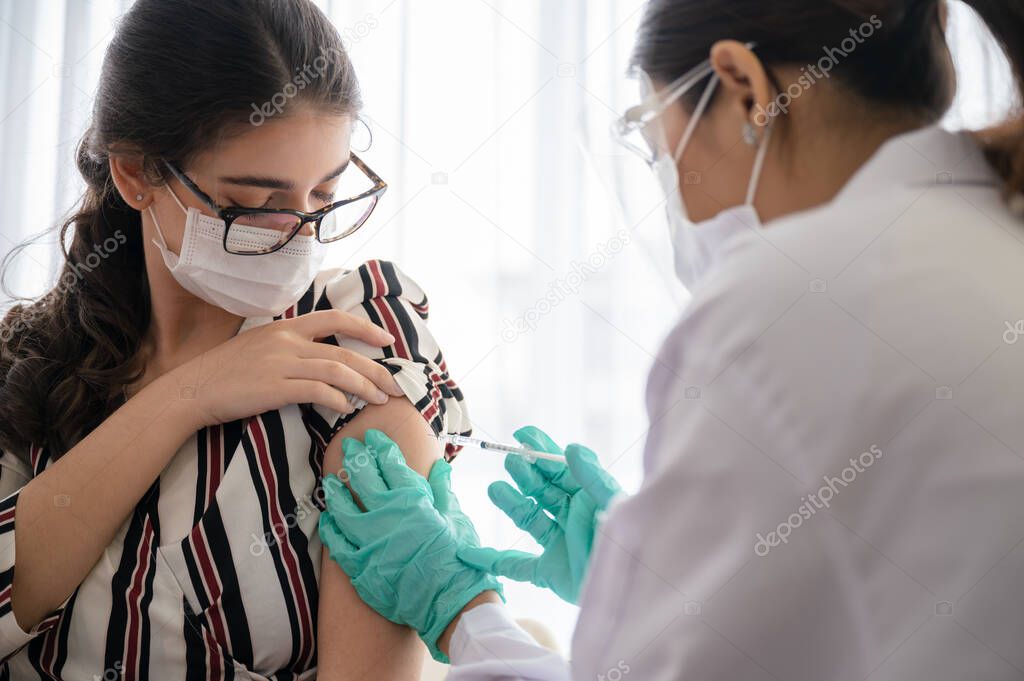 Female doctor or nurse wearing a mask, glove, and face shield are injecting the coronavirus 19 vaccine on shoulder of the woman to immunize in laboratory. Concept of preventing the spread of COVID-19.