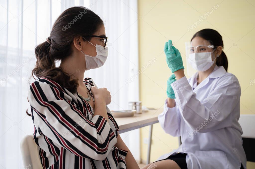 Female doctor or nurse wearing a mask, gloves and visor holds a syringe and coronavirus 19 vaccine, with a female sitting in laboratory waiting for injections vaccine. Concept of preventing COVID-19.