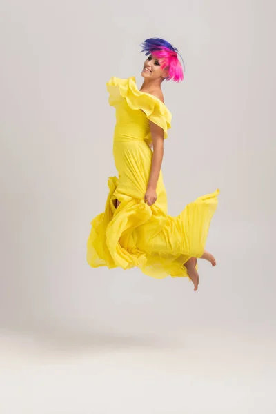 Retrato completo de uma mulher com cabelo colorido brilhante, corte de cabelo azul e rosa. Coloração do cabelo, menina com cabelo curto vestindo em vestido amarelo longo com ombros abertos pulando em um cyclorama leve — Fotografia de Stock