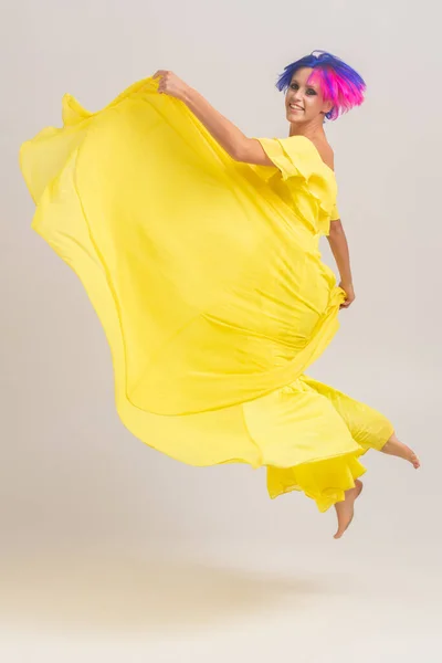 Retrato completo de uma mulher com cabelo colorido brilhante, corte de cabelo azul e rosa. Coloração do cabelo, menina com cabelo curto vestindo em vestido amarelo longo com ombros abertos pulando em um cyclorama leve — Fotografia de Stock