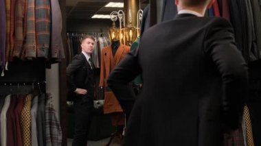 Aynanın önünde duran bir adamı vurmak, kollarını düzeltmek ve mankenlerin arka planına karşı lüks bir erkek giyim mağazasında ceketini iliklemek...