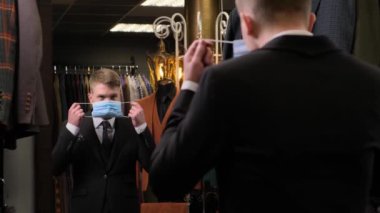 Genç adam aynanın önünde koruyucu bir maske takıyor. Lüks bir erkek giyim mağazasında. Ceketlerin arka planına karşı.