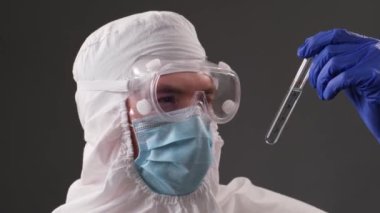 Koruyucu tıbbi kıyafetli doktor elinde bir ampul koronavirüs aşısı tutuyor ve kameraya bakıyor. Salgın ve aşı konsepti. Laboratuvar testi. Klinik araştırması