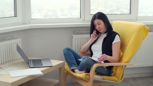 部屋のアームチェアに腰を下ろしてノートパソコンの電話や書類を使っている女性です。自宅で遠隔オンライン作業。ブルネットビジネスの女性はインターネット上でフリーランスをやっている — ストック動画