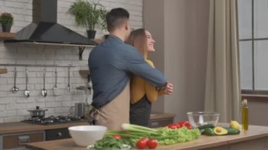 Genç mutlu koca, gülümseyen karısına sarıldı. Mutfakta salata için sebze doğradı. Sevecen çift birlikte yemek pişirdi. Romantik yemek için sağlıklı bir yemek hazırla.