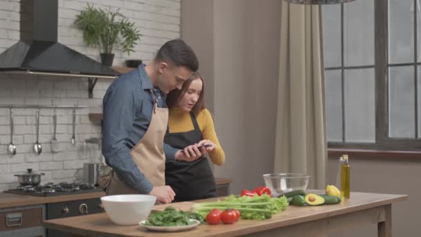Couple regarde la recette sur le téléphone dans la cuisine pour la salade biologique. Mari et femme cuisine recette nourriture. Joyeux mode de vie sain ensemble. Famille recherchant un repas en ligne. — Video
