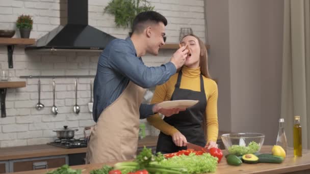 Pareja en el amor preparando comida vegetariana en la cocina casera, marido alimentar a su amada esposa con pizza hablando disfrutar de la fecha de cocción juntos. Relación romántica, concepto de plato saludable — Vídeos de Stock