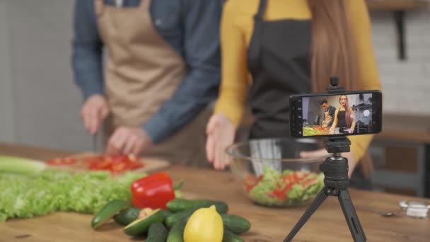 Pareja joven cocinando juntos y grabando video blog de comida en cámara en la cocina en casa. enfoque en la pantalla del teléfono — Vídeos de Stock