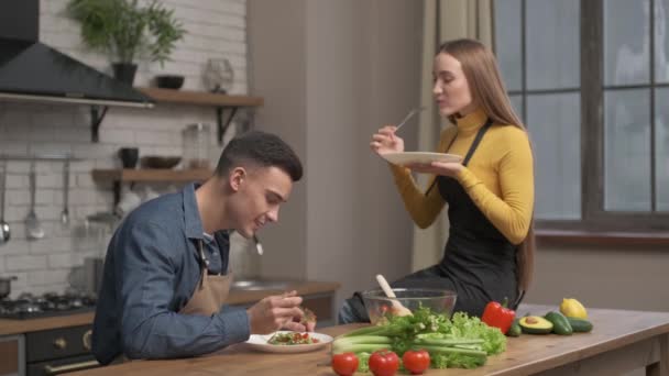 Heureux couple amoureux cuisiné dîner dans une cuisine à aire ouverte, manger de la salade qu'ils se sont préparés dans la cuisine. Une femme est assise sur la table à côté. ils se sont réjouis — Video