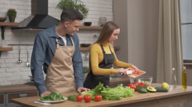 Mutlu çift evde birlikte yemek pişirmekten zevk alıyor. Akşam yemeği için sebze doğrayan ve yanında duran erkek arkadaşıyla konuşan genç bir kadının yan görüntüsü. Neşeli adam ve aşık kadın akşam yemeği için salata yapıyorlar.