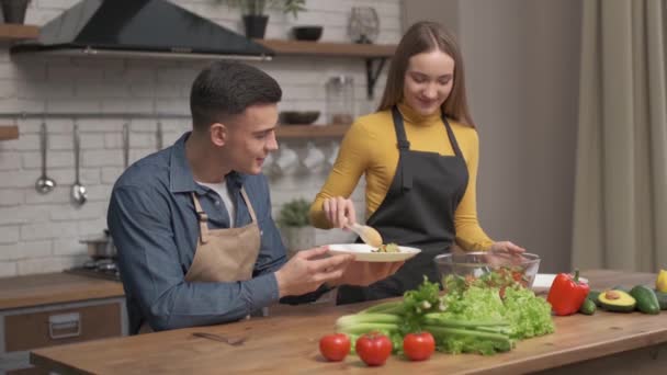 用大玻璃碗把沙拉倒进盘子里 一对恋人在家里厨房做晚饭快乐 健康美味的食物 — 图库视频影像