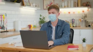 Yüzünde tıbbi maske olan beyaz bir adam evde bilgisayarın başında çalışıyor. Adam öksürüyor.